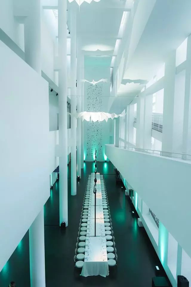 Una mesa larga preparada para cenar en el atrio blanco del MACBA, iluminada en turquesa